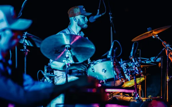 Miglio behind the cymbals in blue light, a country hat blurred in the foreground