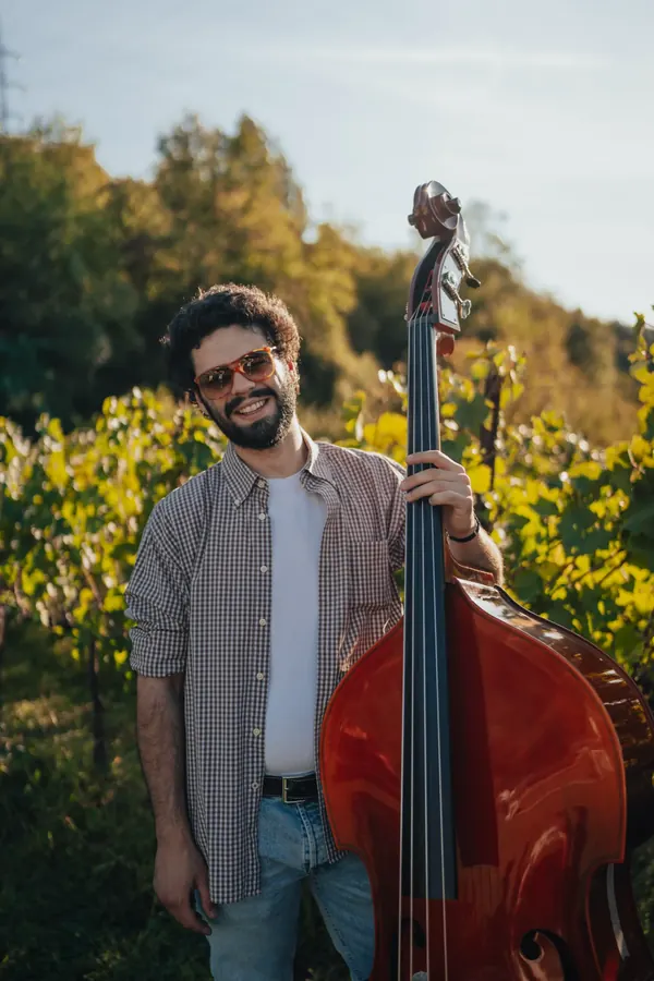 Richi smiling in sunglasses with his double bass among the vines at sunset
