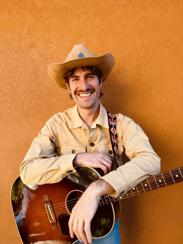 Michele &ldquo;Mike&rdquo; Frosio con cappello da cowboy e chitarra acustica, davanti a un muro arancio