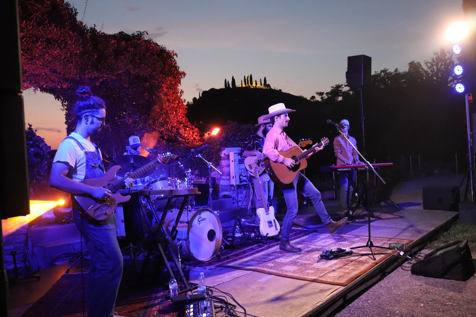 Hot Joe & The CoffeeKillers dal vivo sulla terrazza al tramonto: chitarre, batteria e tastiere sul palco, con la collina e i cipressi sullo sfondo